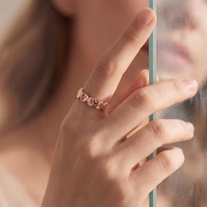 May include: A rose gold ring with a repeating heart design. The ring is worn on a hand, with the focus on the jewelry. The ring is a delicate piece of jewelry.