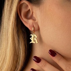 Può includere: Un orecchino a cerchio dorato con un ciondolo pendente a forma di lettera R, realizzato in un carattere gotico stilizzato. L'orecchino è realizzato in metallo color oro lucido e presenta un semplice design a cerchio circolare con un'iniziale.