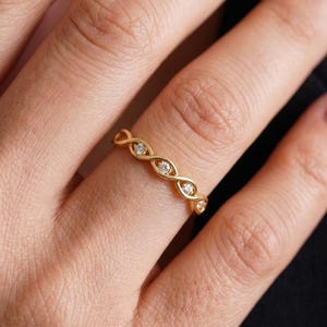 May include: A gold ring featuring a twisted, infinity-style band design. The metal band is accented with three small, clear, round-cut stones set within the center of the intertwined sections, creating a delicate and continuous metallic pattern.