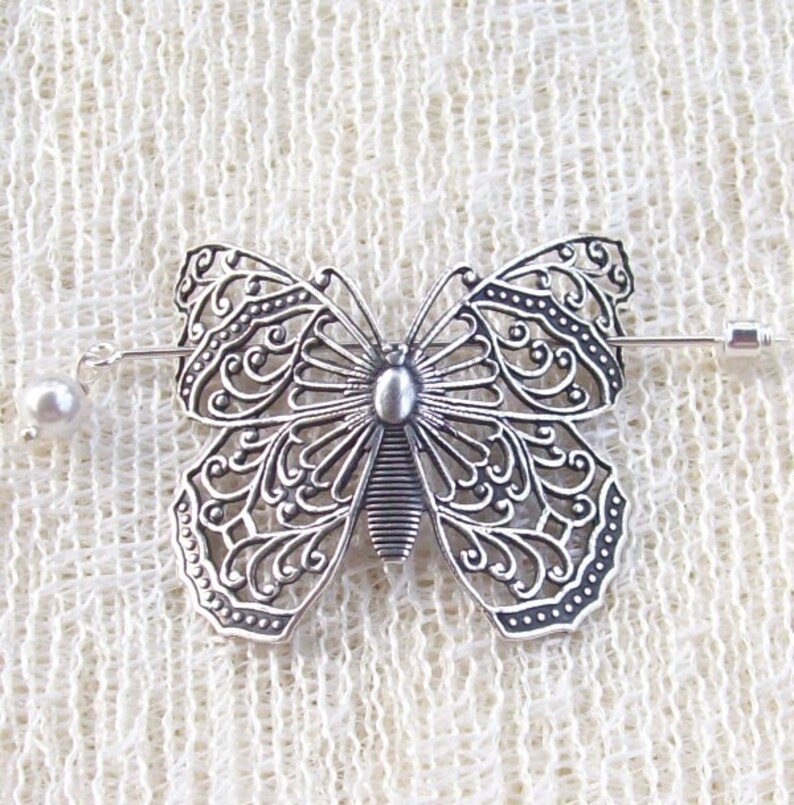 May include: Silver filigree butterfly brooch with a pearl accent. The brooch is attached to a silver pin.