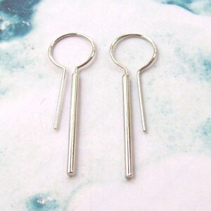 May include: A pair of minimalist silver earrings. Each earring features a circular top with a straight, elongated bar pendant. The earrings are displayed against a light blue and white marbled background.