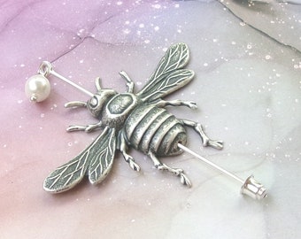 May include: A silver bee brooch with a pearl accent. The bee is detailed with textured wings and a segmented body. The brooch is attached to a silver pin.