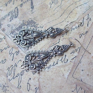 May include: A pair of silver dangle earrings with intricate filigree designs. The earrings are made of metal and have a vintage style.