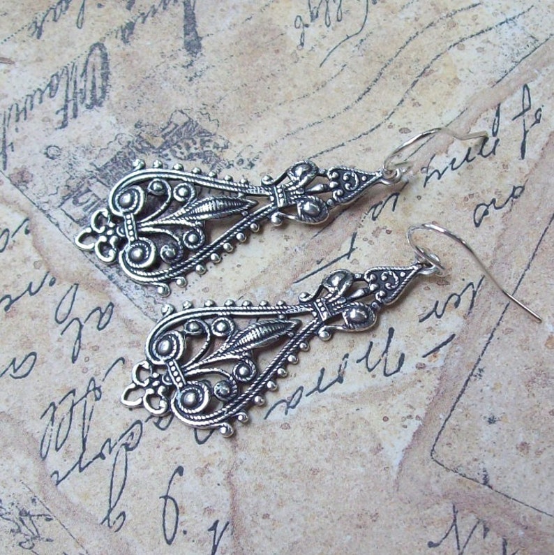 May include: A pair of silver dangle earrings with intricate filigree designs. The earrings are made of metal and have a vintage style.