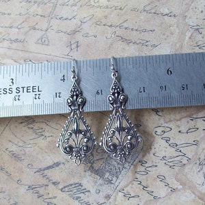 May include: A pair of silver dangle earrings with a filigree design. The earrings are approximately 2 inches long and are measured against a ruler.
