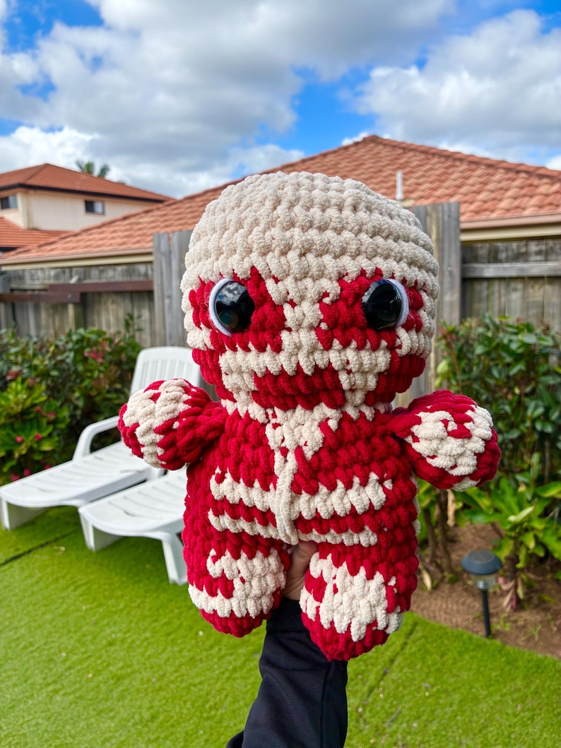 Puede incluir: Un peluche hecho a mano con ganchillo en colores rojo y crema. El juguete tiene ojos grandes y saltones y un dise&ntilde;o detallado que se asemeja a un personaje de una serie de anime. El peluche se sostiene sobre un fondo de hierba verde y un barrio residencial.