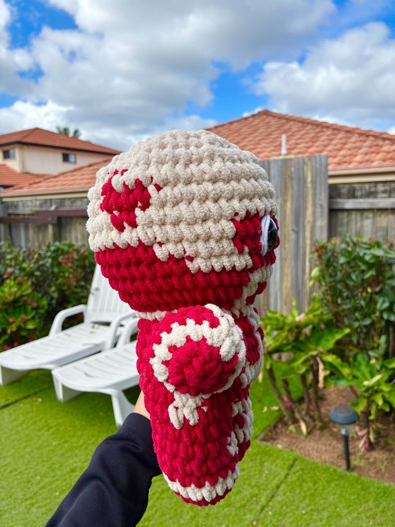 Puede incluir: Una mu&ntilde;eca de peluche hecha a mano, con cabeza y cuerpo redondos. La mu&ntilde;eca es principalmente roja con detalles blancos, incluido un patr&oacute;n en la cabeza y el cuerpo. Tiene ojos grandes, negros y blancos. La mu&ntilde;eca est&aacute; hecha de un hilo suave y texturizado.