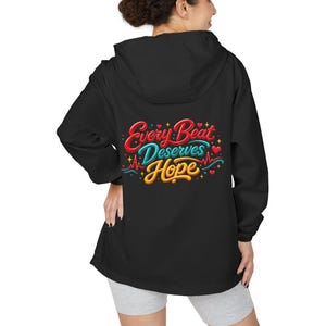 Könnte beinhalten: Eine schwarze Kapuzenjacke mit einer Grafik auf der Rückseite, die den Text Every Beat Deserves Hope in roter, türkisfarbener und gelber Schreibschrift zeigt. Das Design enthält eine rote EKG-Herzrhythmuslinie, kleine rote Herzen und gelbe Sterne.