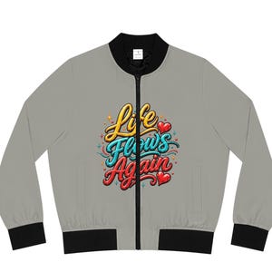 Könnte beinhalten: Eine hellgraue Bomberjacke mit schwarzem Rippstrickkragen, Bündchen und Saum. Auf der Vorderseite befindet sich ein zentrales Motiv mit dem Schriftzug "Life Flows Again" in stilisierter gelber, blauer und roter Schrift, akzentuiert durch kleine rote Herzen.