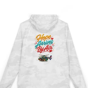 Könnte beinhalten: Eine weiße Kapuzen-Windjacke mit Camouflage-Muster, die auf der Rückseite eine Grafik mit dem Text Hope Arrives By Air in gelber, türkiser und roter Schreibschrift sowie eine bunte Hubschrauber-Illustration und kleine rote Herzen zeigt.