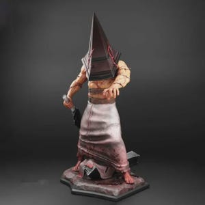 Pode incluir: Estatueta detalhada de um personagem com cabeça em forma de pirâmide, segurando uma faca grande. A figura é retratada com um torso musculoso, envolto em bandagens e usando um avental rasgado. A base é texturizada.