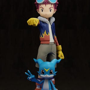 May include: A figurine set featuring two characters from the Digimon anime series. The first character is a young boy with red hair, goggles, and a blue jacket with flame details. The second character is a blue creature with large ears and a peace sign.