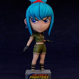 Puede incluir: Figura de un personaje femenino con cabello azul y una diadema roja, vestida con un atuendo verde de estilo militar y guantes negros. La figura está sobre una base que dice "THE KING OF FIGHTERS".