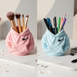 May include: Two desk tidies in the shape of hoodies, one pink and one light blue, each with a black swoosh logo. The pink one holds makeup brushes, the blue one pens and pencils. Both are on a white surface.