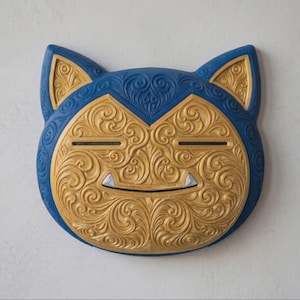 May include: A wall-mounted mask shaped like a cat head with pointed ears. The mask features a deep blue outer border and a central gold face, both decorated with intricate, swirling, embossed patterns. Two small white fangs are visible at the mouth.
