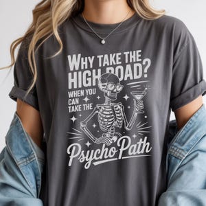 May include: Dark gray t-shirt with the text "Why take the high road? When you can take the Psycho Path." A skeleton wearing sunglasses and holding a martini glass is featured in the graphic. The shirt has rolled-up sleeves.