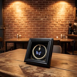 May include: A square black frame containing a circular arrangement of a white snake skeleton with a small blue butterfly positioned in the centre, set against a black textured background with an embossed floral pattern.