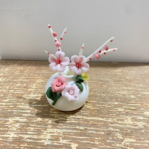 Pode incluir: Um pequeno vaso de cerâmica branco e redondo decorado com rosas cor-de-rosa esculpidas e folhas verdes. Dois ramos brancos com pequenos botões cor-de-rosa e duas flores cor-de-rosa maiores de cinco pétalas saem da abertura.