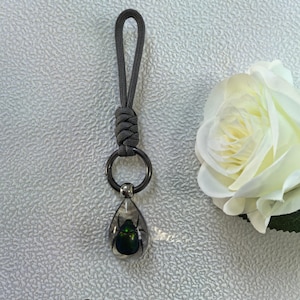 Puede incluir: Un llavero de paracord gris carbón con un lazo anudado unido a un anillo de metal oscuro. Un colgante de resina transparente en forma de lágrima cuelga del anillo y contiene un escarabajo verde iridiscente. El texto Charcoal gray es visible.