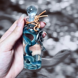 May include: A clear glass bottle with a spherical stopper containing a blue snake figurine submerged in liquid. A tan leather cord is tied around the neck of the bottle, featuring a silver-toned metal charm shaped like an Egyptian ankh symbol.