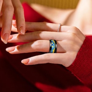 May include: A black ring with a starry night design in yellow and blue. The ring is on a finger, and another ring with small clear stones is also visible. The background is a red sweater.