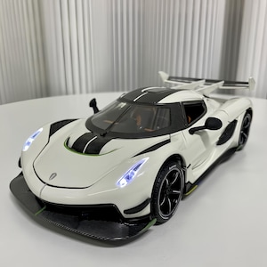 May include: A white scale model sports car featuring a black carbon fibre bonnet stripe, a large rear wing, and black wheels. The vehicle has illuminated LED headlights, a low-profile aerodynamic front splitter, and visible green accent detailing.