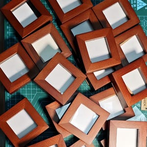May include: A collection of square wooden picture frames with a medium brown finish and a visible wood grain texture. Each frame features a white centre insert and a beveled edge design. The frames are arranged in a scattered, overlapping pattern.