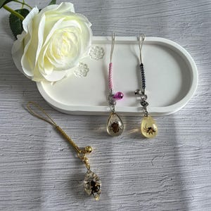 Puede incluir: Tres colgantes para teléfono con piezas de resina transparente que contienen insectos. Uno tiene forma de hoja con cordón dorado, mientras que dos tienen forma de lágrima con cordones trenzados rosa y negro, decorados con una pequeña campana.