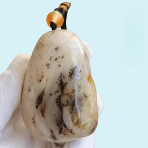 May include: A smooth, teardrop-shaped pendant made of off-white dendritic agate with dark, fern-like inclusions scattered across its surface. The pendant is suspended by a dark cord featuring a small, round, translucent orange bead at the top.