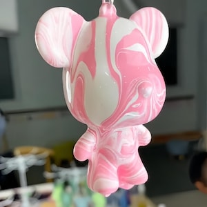 Op de afbeelding: Een berenvormige sleutelhanger met een glanzend, gemarmerd wervelpatroon in wit en roze. De hanger is bevestigd aan een zwarte plastic clip en een zilveren metalen ring. De tekst "Purely handmade on-site casting" is zichtbaar.