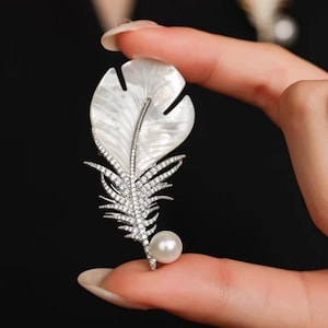 May include: A feather-shaped brooch with a pearlescent white top and silver-coloured stem and accents. The brooch is decorated with small, sparkling crystals and a single, round pearl at the bottom. The background is black.
