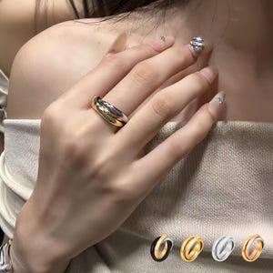 Pode incluir: Um anel dourado e prateado com um design arredondado. O anel é mostrado no dedo de uma pessoa. Anéis adicionais em preto, dourado, prateado e ouro rosa são mostrados na parte inferior da imagem.