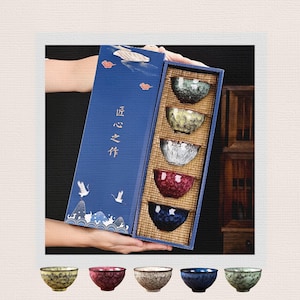 Puede incluir: Un juego de cinco cuencos de té de cerámica en una caja de regalo rectangular azul. Los cuencos presentan variados patrones de esmalte craquelado en verde, amarillo, blanco, rojo y azul. La tapa muestra grullas y caracteres chinos.
