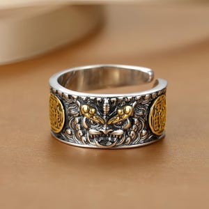 Op de afbeelding: Verstelbare ring in zilverkleur met een gedetailleerd ontwerp. De ring heeft een centraal gezicht met goudkleurige accenten en twee cirkelvormige goudkleurige emblemen aan weerszijden. Het ontwerp van de ring is ingewikkeld en gedetailleerd.