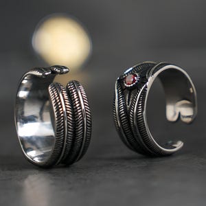May include: Two silver-toned open rings featuring a detailed feather-like texture. One ring is plain, while the other is accented with a small, round, deep red gemstone set within a dark, recessed center detail on the outer band surface.