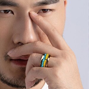 May include: Gold-toned ring with a colorful design. The ring features a band with blue, yellow, and black accents. The ring is worn on a finger.