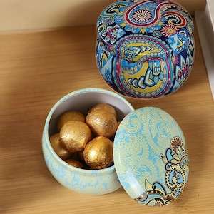 May include: Two round metal tins featuring intricate paisley patterns. One tin is closed, displaying vibrant blue, yellow, and red colors. The other tin is open, revealing a light blue exterior with gold-colored spherical items wrapped in gold foil.