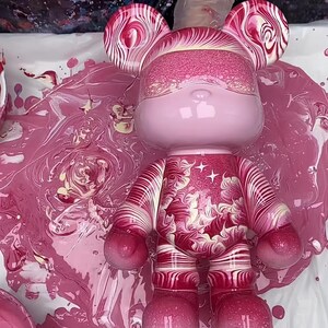 May include: A bear-shaped figurine featuring a glossy light pink head with a textured pink band across the eyes. The torso and limbs display a swirling marbled pattern in shades of deep magenta, white, and pink, accented by two small white stars.