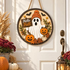 Peut inclure: Une plaque de porte ronde en bois représentant un fantôme blanc portant un bonnet orange, tenant un panneau avec l'inscription Hello Fall. Le décor inclut des citrouilles orange, des petites citrouilles blanches, des feuilles et des chauves-souris.
