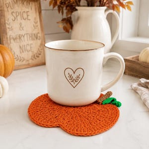 May include: A white ceramic mug with a brown heart and leaf outline design sits on a bright orange crocheted pumpkin-shaped coaster. The coaster features a small brown stem and a green leaf detail, providing a textured base for the mug.