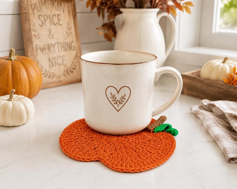 May include: A white ceramic mug with a brown heart and leaf outline design sits on a bright orange crocheted pumpkin-shaped coaster. The coaster features a small brown stem and a green leaf detail, providing a textured base for the mug.