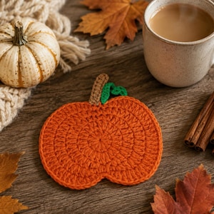 May include: A round, orange crochet coaster shaped like a pumpkin, featuring a small brown stem and a green leaf detail at the top. The coaster is crafted with a circular stitch pattern and is displayed as a flat, decorative textile item.
