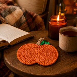 May include: A round, orange crocheted coaster shaped like a pumpkin, featuring a small brown stem and a single green leaf attached to the top. The coaster is crafted with a textured, circular stitch pattern and is laid flat on a wooden surface.