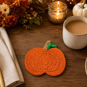 May include: A round, orange, crocheted pumpkin coaster featuring a textured surface, a small tan stem, and a single green leaf attached at the top. The coaster is crafted from yarn with visible circular stitching patterns throughout the design.