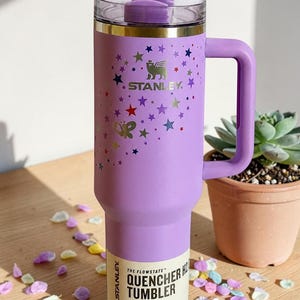 May include: A purple Stanley Flowstate Quencher tumbler with a handle, decorated with colorful stars and a butterfly. The tumbler has a clear lid and a label that reads "Quencher H2.0 Tumbler". It holds 40 oz (1.18 L).