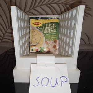 Può includere: Un contenitore di stoccaggio bianco aperto con un pacchetto di zuppa Waldpilz Cremesuppe. La confezione della zuppa è gialla e verde, con un'immagine della zuppa in una ciotola. Una nota scritta a mano sotto dice "SOUP".