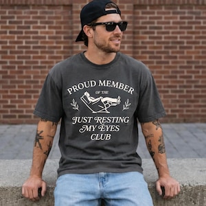 May include: Dark gray t-shirt with the text "PROUD MEMBER of the JUST RESTING MY EYES CLUB" in white script. The shirt features a graphic of a person reclining in a chair. The shirt is worn by a person wearing sunglasses and a black cap.