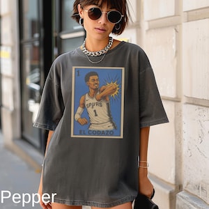 May include: A charcoal gray short-sleeve t-shirt featuring a graphic print of a basketball player in a white Spurs jersey with the number 1. The design resembles a playing card with the text 1 and EL CODAZO printed in a blue rectangular frame.
