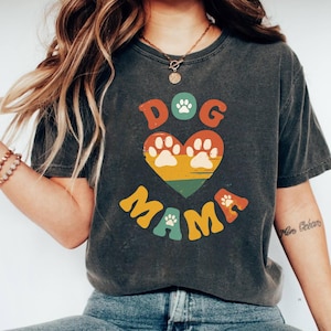 May include: Dark gray t-shirt with the words "DOG MAMA" in colorful, retro-style lettering. The design includes a heart-shaped graphic with paw prints and a rainbow color scheme. The shirt has a vintage, worn look.