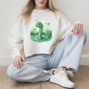 May include: White sweatshirt featuring a green cartoon illustration of a Loch Ness Monster in water, with shamrocks. The design is centered on the front of the long-sleeved top. The sweatshirt is paired with light blue jeans and white sneakers.